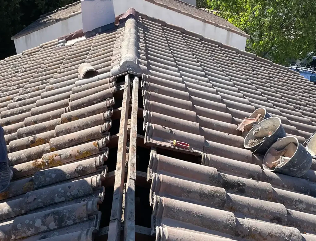 Un toit de tuiles dont quelques tuiles ont été enlevées le long du faîtage, révélant un vide. Des outils et des chaussures de travail sont visibles sur le toit, ce qui témoigne d'un changement continu du patrimoine. Des arbres et une partie de la façade de la maison apparaissent à l'arrière-plan.