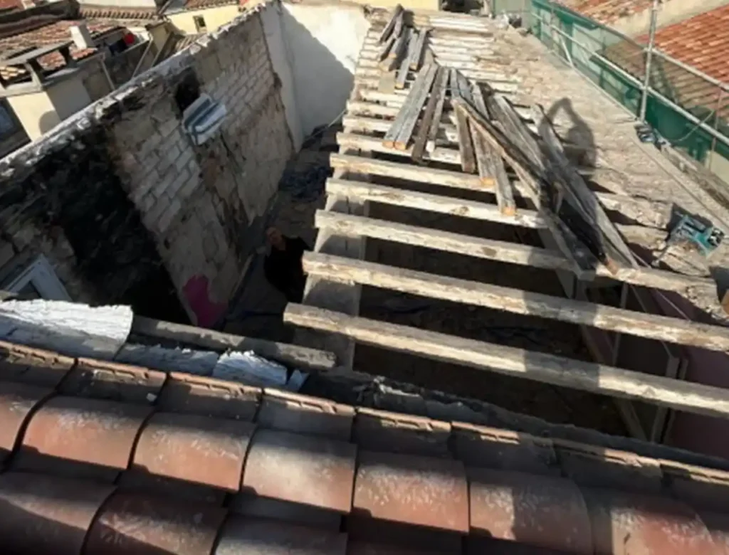 Vue d'un bâtiment partiellement démoli lors d'une dépose toiture, avec des poutres de toit et des murs en bois apparents ; des piles de planches en bois reposent sur les poutres, entourées de matériaux de construction et de débris en dessous.