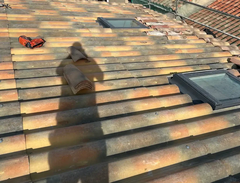 Un toit en tuiles éclairé par le soleil, avec deux fenêtres, des tuiles détachées et un outil rouge sans fil reposant sur la surface, fait penser à une création de toiture neuve. L'ombre d'une personne est visible sur le toit, ce qui suggère que des travaux sont en cours.
