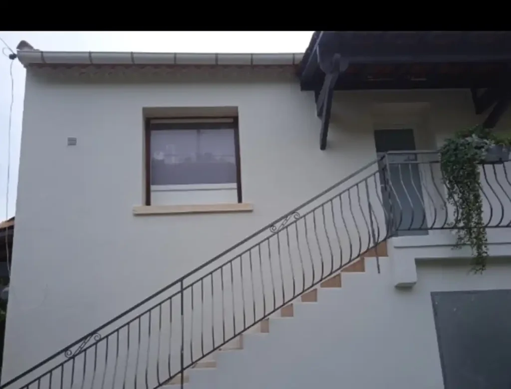 Un bâtiment de couleur claire avec une peinture de façade fraîchement appliquée présente un escalier extérieur menant à une porte à l'étage supérieur. Une fenêtre se trouve à gauche de l'escalier et une plante est suspendue à la balustrade près de l'entrée.