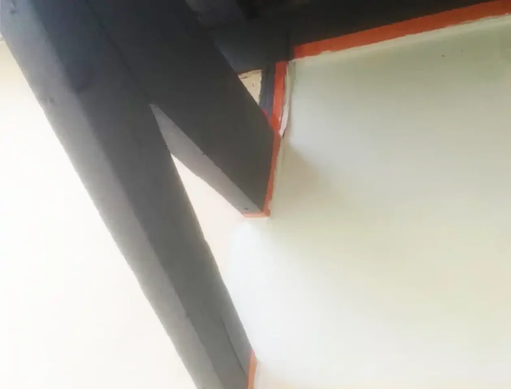 Vue rapprochée d'un mur blanc rencontrant une poutre en bois foncé, avec une peinture de façade rouge inégale et désordonnée le long des bords où la poutre et le mur se croisent.