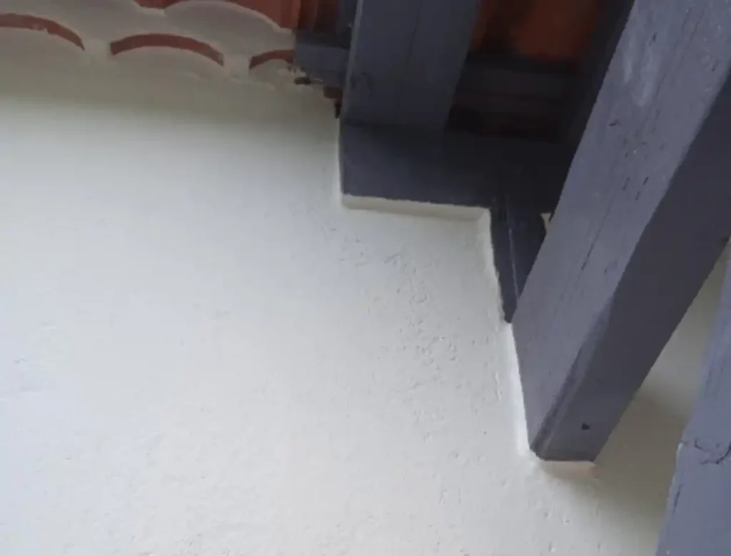 Gros plan d'un mur enduit de blanc avec peinture de façade rencontrant un plafond en poutres de bois gris foncé, avec peinture écaillée et toiture apparente dans le coin supérieur gauche.